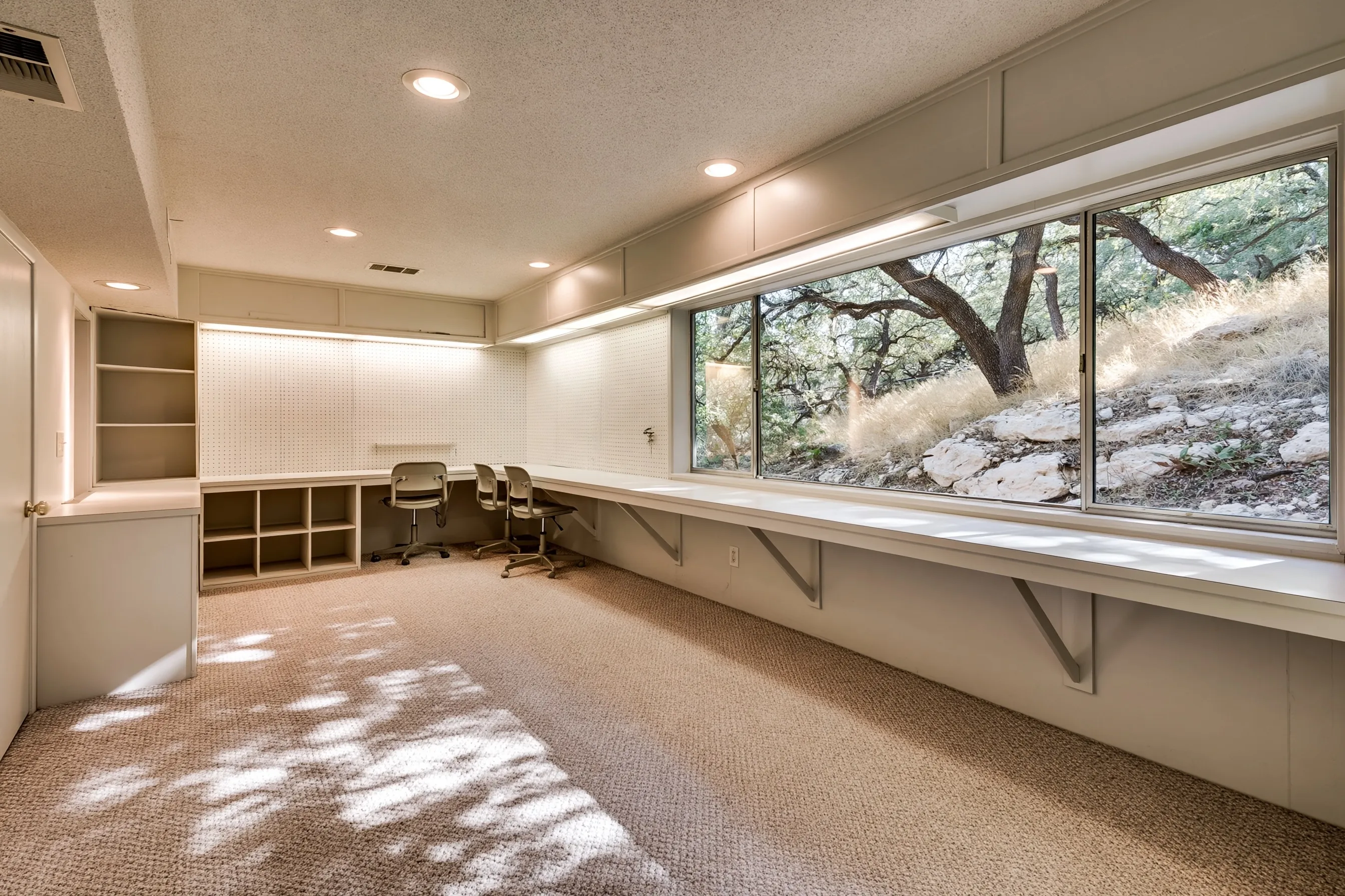 Walkout-level office with a long L-shaped desk counter on brackets, pegboard, cubbies, three chairs and a wide window onto a rocky hillside with oaks