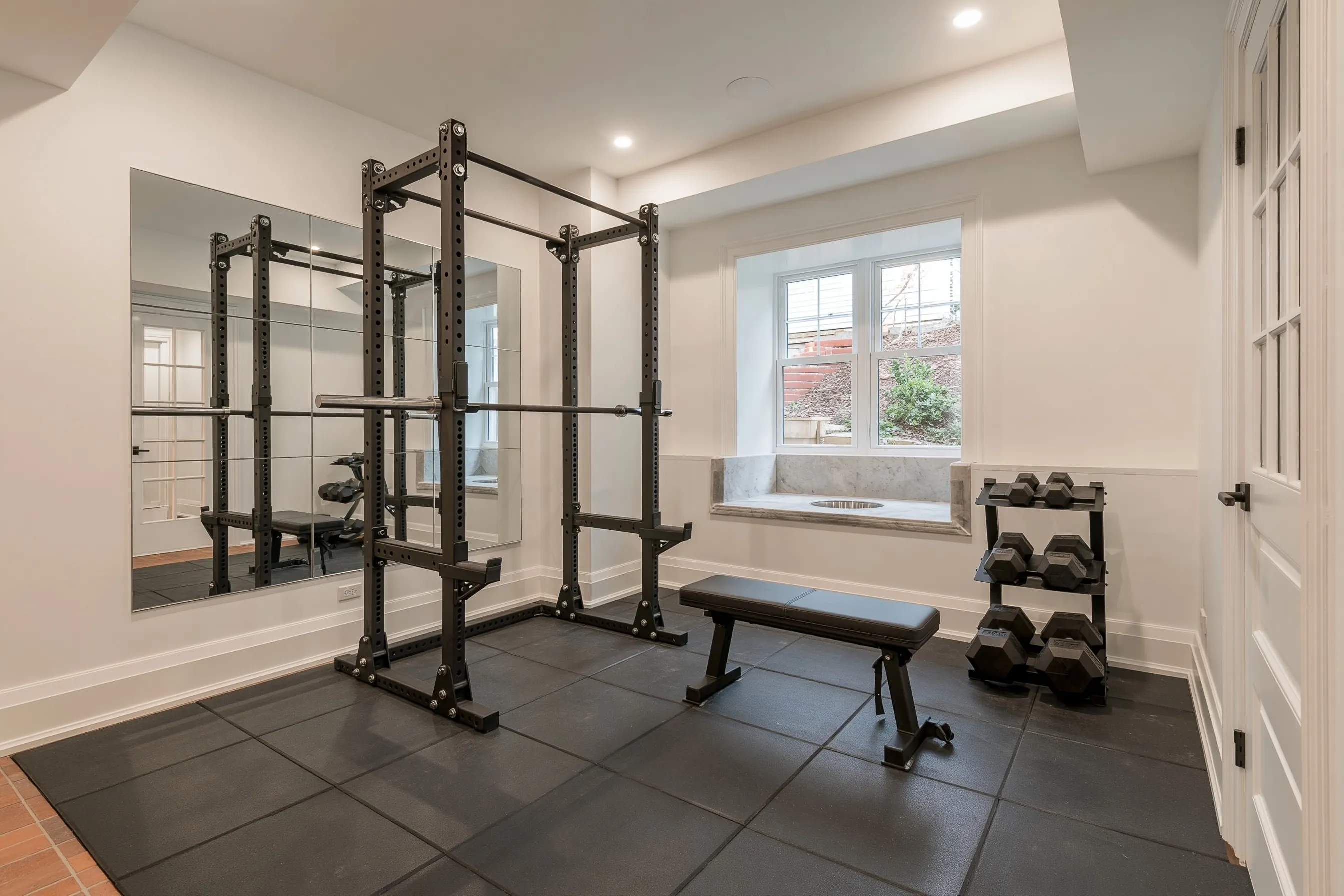 Basement home gym with black rubber floor tiles, a mirror wall, a squat rack, a bench, dumbbells and a window in a deep well